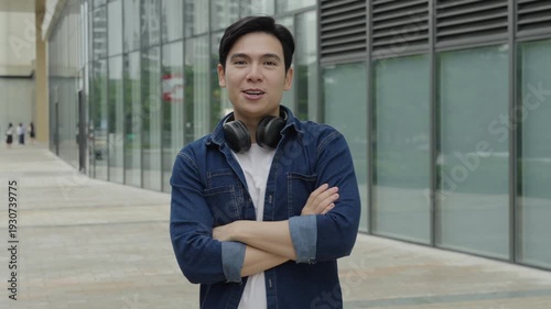 full body shot of a young Asian man crossing his arms with a confident gaze in a urban landscape. The man wears a casual denim shirt and has headphones around his neck