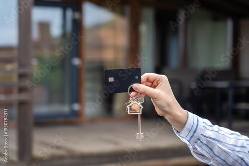 Hand holding keys with plastic credit card in front of new house. Moving relocation, mortgage, property, real estate concept. Buying or renting