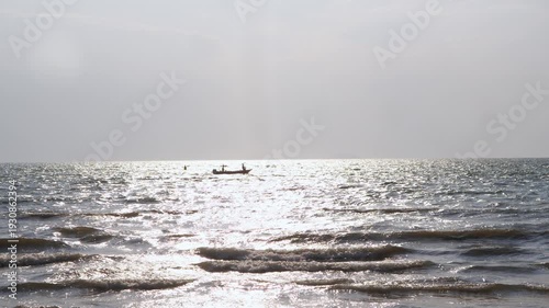 Small motorboat moves across shimmering sunlit sea surface far from shore. Gentle waves roll in foreground under bright hazy sky.