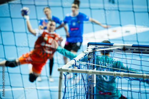 View through the handball net showing a blurred player in an orange jersey jumping to score a goal during a professional competition.