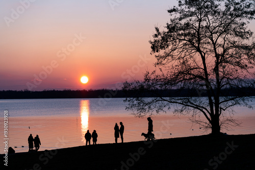 Sunset at Cospuden Lake, Markkleeberg near Leipzig, people and a tree as silhouettes against a colorful sky, tranquil lakeside evening scene.