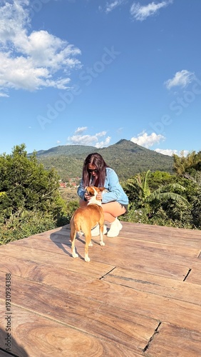 A young woman enjoying a peaceful outdoor moment with her dog on a wooden deck overlooking lush green mountains under a bright blue sky