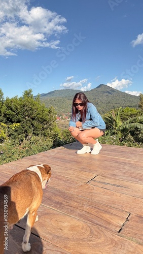 A young woman enjoying a peaceful outdoor moment with her dog on a wooden deck overlooking lush green mountains under a bright blue sky