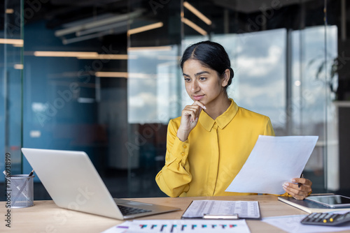 Serious and thoughtful young Indian woman sitting at her desk. holding documents and looking at laptop screen