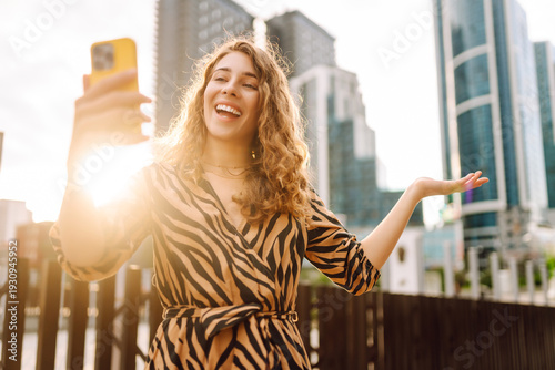Φωτογραφία A smiling woman with phone in hand is blogging or videoconferencing against backdrop of metropolis