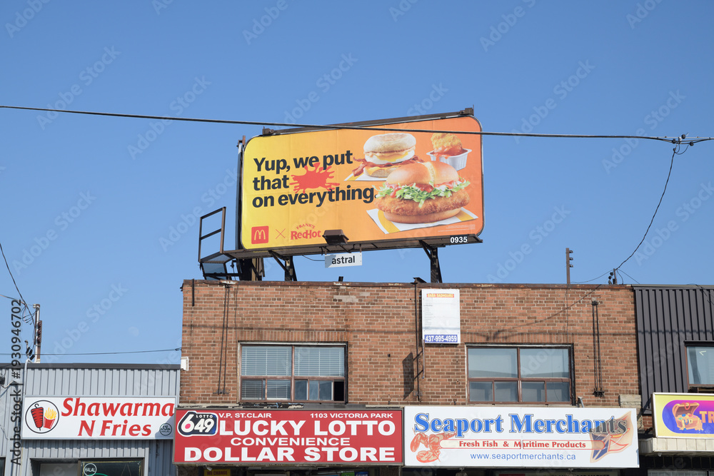 Naklejka premium rooftop billboard of McDonald's Canada collab with Frank's Red Hot Sauce and shop signs (Shawarma N' Fries, Lucky Lotto Convenience Dollar Store, Seaport Merchants) Victoria Park Av, Toronto