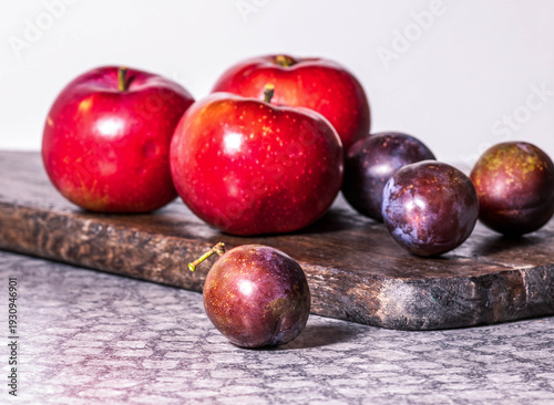 Red apples and purple plums on wooden board against marble surface