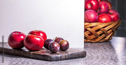 Fresh red apples and plums on wooden cutting board with basket