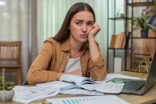 Young woman at home table holds head stressed while unpaid bills cover surface beside laptop and calculator. Caucasian girl feels overwhelmed and searches for solutions to manage urgent debt pressure.
