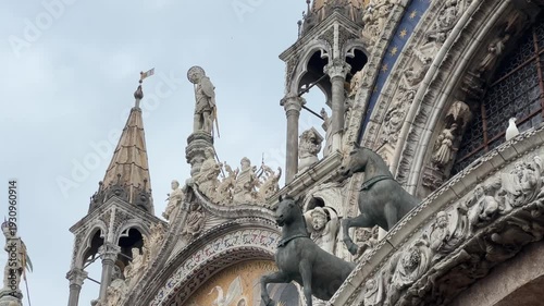 A close-up of the stunning St. Mark’s Square (Piazza San Marco) in Venice, highlighting the architectural details of the basilica’s facade. A seagull flies past the golden mosaics and bronze horses.