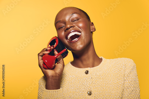 Slika na platnu Joyful African woman with dark skin laughing and holding red sunglasses near her face