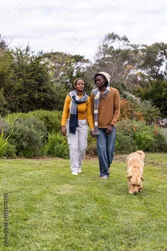 Obraz African American couple holding hands walking in park wearing sweaters and scarves with tan dog