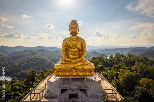 Breathtaking aerial panorama of the large golden Phra Phuttha Lokanat Satsada meditation Buddha statue at Wat Wang Wiwekaram overlooking winding river, lush forest, and layered mountain landscape in S
