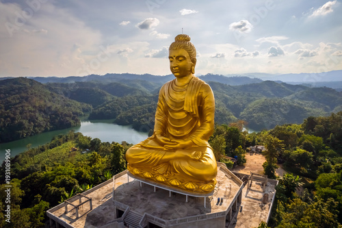 Breathtaking aerial panorama of the large golden Phra Phuttha Lokanat Satsada meditation Buddha statue at Wat Wang Wiwekaram overlooking winding river, lush forest, and layered mountain landscape in S