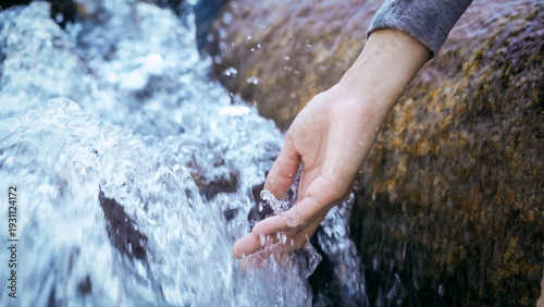Close-up of a human hand reaching into a fast-flowing clear mountain stream beside a mossy rock, splashing water, outdoor nature scene.