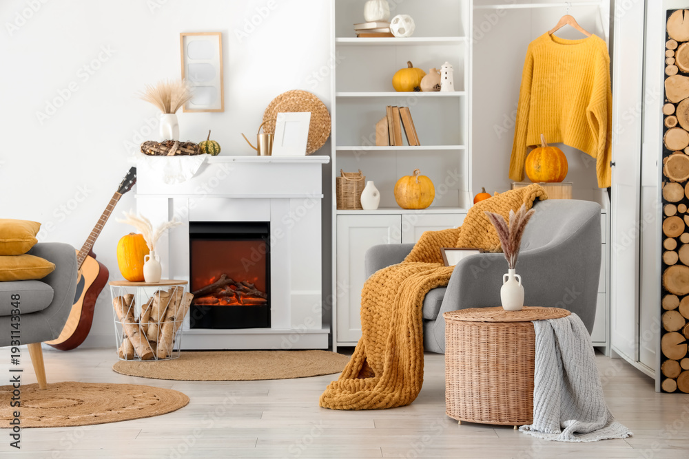 Fototapeta premium Interior of living room with armchairs, coffee tables, guitar, autumn home decor on fireplace and pumpkins in shelving unit near white wall