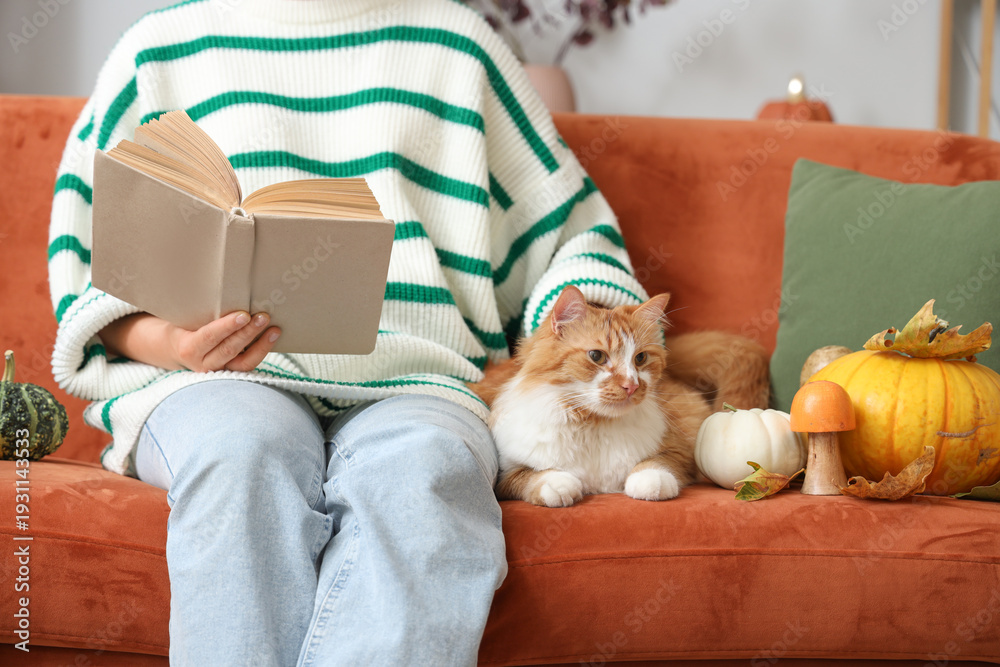 Fototapeta premium Young woman with cute cat reading book at home on autumn day