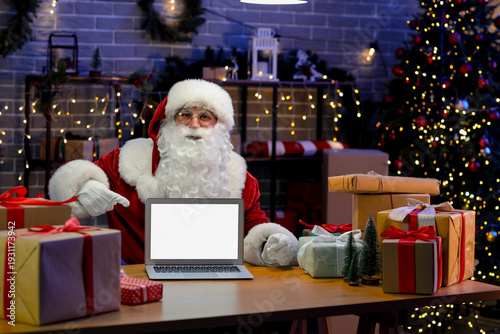Santa Claus with gift boxes pointing at blank laptop in post office