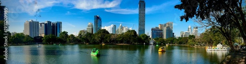 Colorful swan paddle boats glide across the calm lake at Lumpini Park in Bangkok, framed by lush trees and a modern skyline under clear blue skies on a bright afternoon.