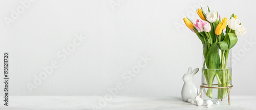 Glass vase with beautiful tulip flowers, Easter eggs and ceramic bunny on light background