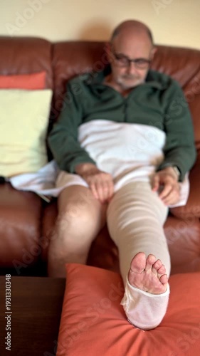 Relaxed elderly man with a bandaged leg resting on the sofa in his home