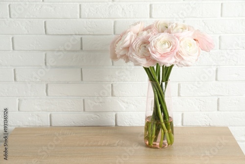 Beautiful ranunculus flowers in glass vase on wooden chest of drawers near white brick wall indoors. Space for text