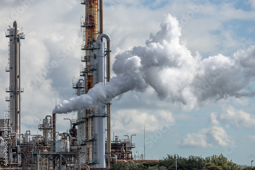 Photos Industrial refinery plant complex emitting dense vapor cloud trail into partially overcast sky from rust-streaked refinery chimney, highlighting emissions, energy production and environmental impact