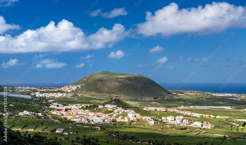 Naklejka premium Scenic sunny view of the Montana de Taco volcanic cone surrounded by green banana plantations and traditional white towns near the ocean in Tenerife, Canary Islands.