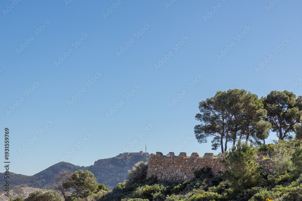 Fototapeta premium The ancient stone wall of Roman-era fortresses in Cartagena, Spain, rises majestically against the backdrop of a rugged mountain, the historic strength and enduring beauty