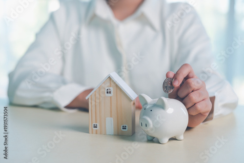 Hand putting coin into piggy bank beside small house model, symbolizing home savings, mortgage planning, real estate investment, property finance, and financial security strategy.