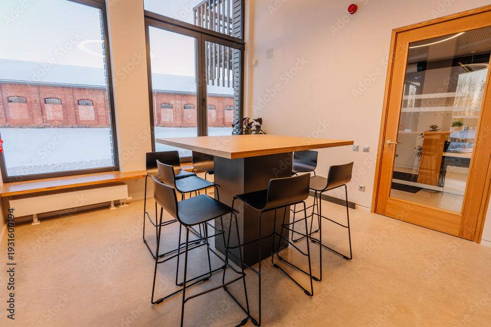 Fototapeta premium Modern interior of a minimalist office kitchen featuring a large wooden table surrounded by black metal stools and large windows with natural light