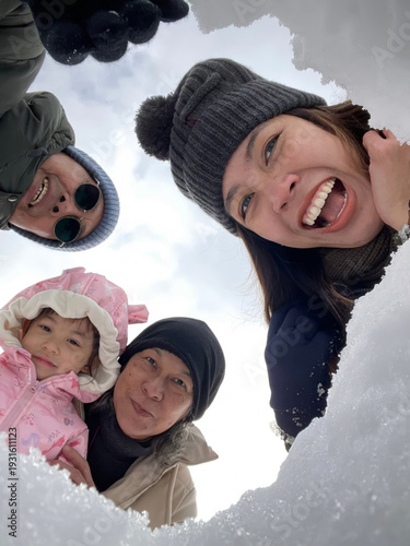 Joyful three-generation Asian family selfie portrait in Japan, featuring grandparents, parents, and a child smiling together through a snow hole, capturing a perfect winter vacation moment.