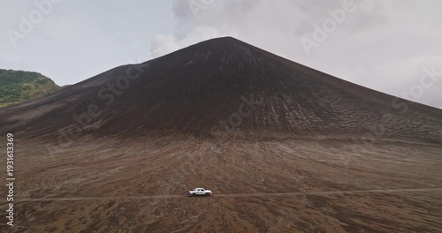 Pickup truck crosses rugged dirt road on vast ash plain toward Mount Yasur volcano under brooding gray skies on remote Tanna Island, Vanuatu raw, dramatic volcanic landscape. Aerial view drone shot