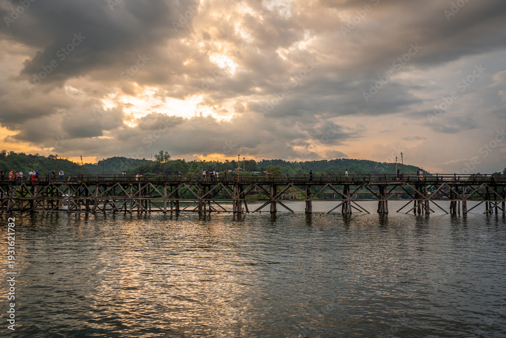 Fototapeta premium Dramatic sunset over wooden Mon Bridge with silhouette traveler crossing calm river in Sangkhlaburi, Kanchanaburi, Thailand