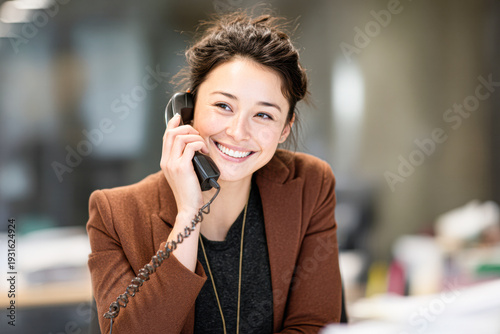 A smiling woman in a brown blazer talks on a corded telephone in a modern office setting.