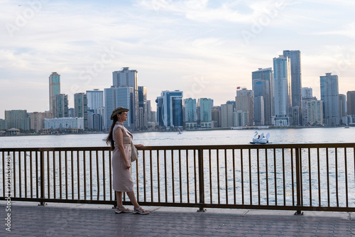 Young woman tourist taking selfie and enjoying sunset view at Sharjah Corniche waterfront with city skyline in background, United Arab Emirates.