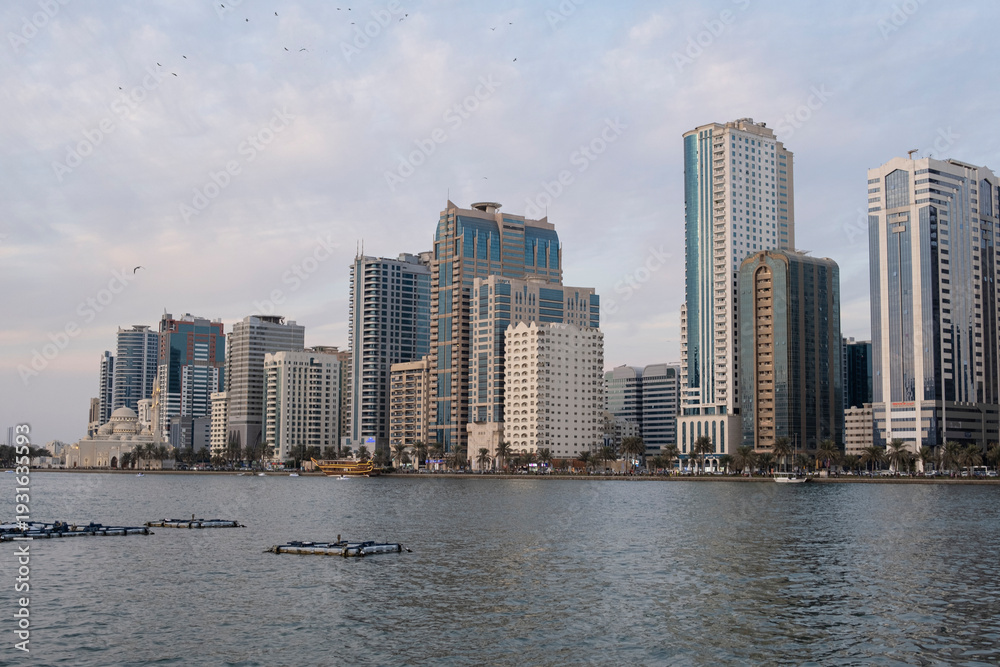 Fototapeta premium 10.12.2025 Sharjah, UAE. Sunset skyline of Sharjah with modern skyscrapers and luxury residential towers along the Corniche waterfront under a golden sky, United Arab Emirates.