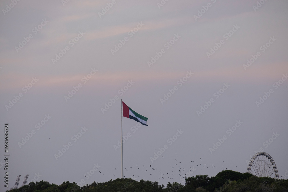 Fototapeta premium National flag of the United Arab Emirates waving on a high flagpole at dusk against the Sharjah city skyline and twilight sky, UAE.