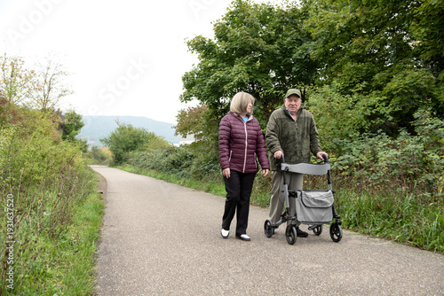 Elderly married couple taking a walk in the park with help of walker to support and help move