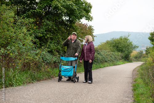 Elderly married couple taking a walk in the park with help of walker to support and help move