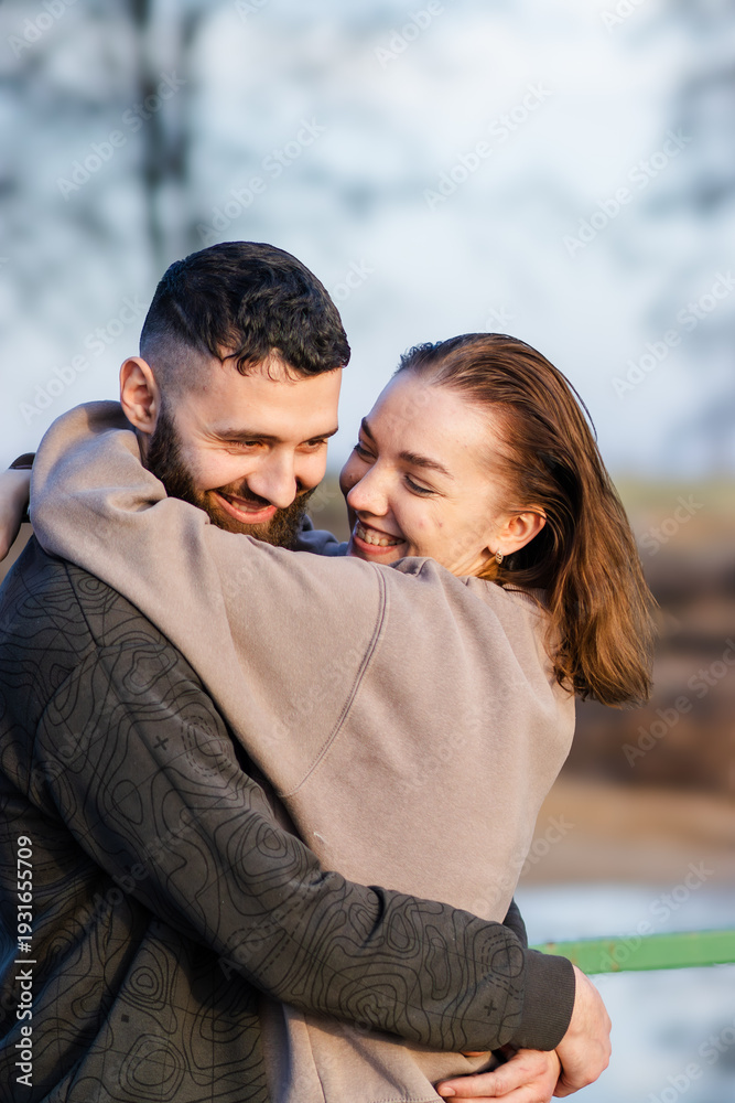 Naklejka premium Young couple embracing outdoors, smiling joyfully while standing close together against a blurred natural background with trees and soft lighting