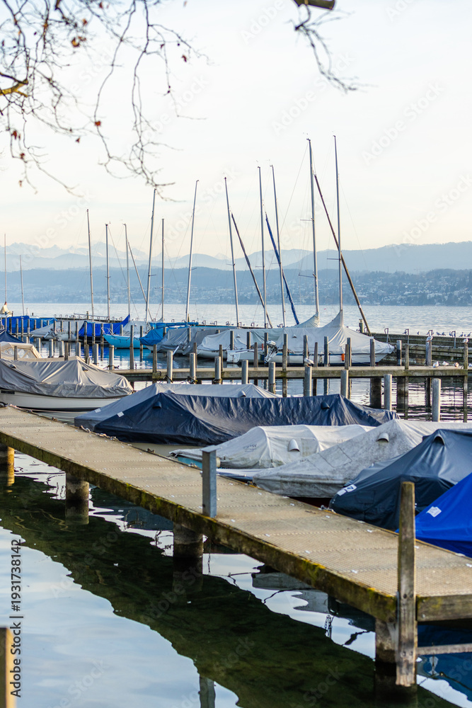 Fototapeta premium Vertical view of a wooden pier with moored sailboats on Lake Zurich (Zürichsee) in Switzerland, featuring the distant snow-capped Swiss Alps under a clear sky.