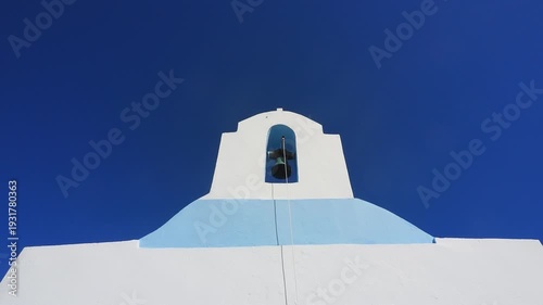 4K footage of a traditional Greek island church bell tower in the Cyclades with a bell rope gently swaying in the wind against a deep blue sky. Clean Mediterranean architecture with minimal compositio
