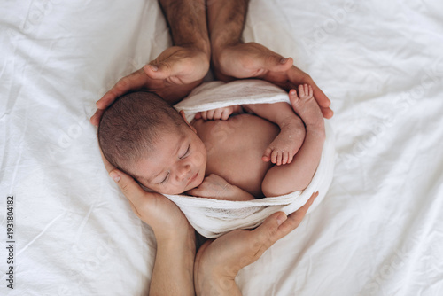 Newborn baby wrapped in soft cloth, cradled gently by adult hands on white bedding