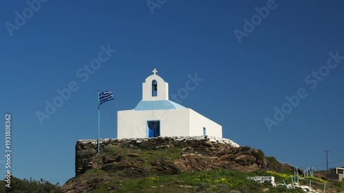 4K footage of a traditional white Greek island chapel overlooking Korissia in Kea, Cyclades. The Greek flag waves in the wind against a deep blue sky. Authentic Mediterranean architecture suitable for