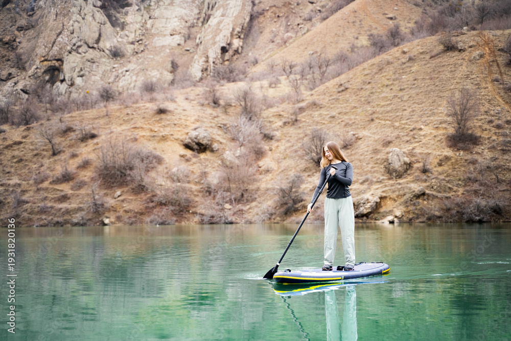Naklejka premium Female on sup board in mountain lake in daylight. Water sport activity and healthy outdoor lifestyle in scenic environment. 