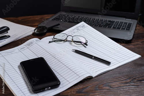 Financial accounting book with a smartphone, glasses, and a pen placed on top. Laptop in the background, business and finance workspace concept.