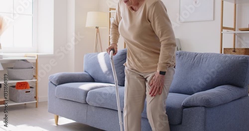 Elderly senior woman with cane, hip pain at home. She sits on sofa in a bright room, gripping her hip, grimacing, needing a crutch or cane. Clear concept pain, injury, limited mobility in retirement.