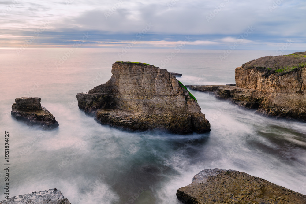 Obraz premium Shark Fin Cove along the California coast featuring dramatic layered rock formations and sea stacks rising from the Pacific Ocean. Long exposure captures smooth water and rugged coastal geology