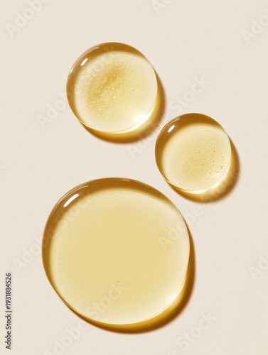 Top view macro image of three glossy cosmetic oil drops on a clean light beige background. The viscous transparent serum has a natural golden tone and smooth texture, suitable for skincare, beauty, sp